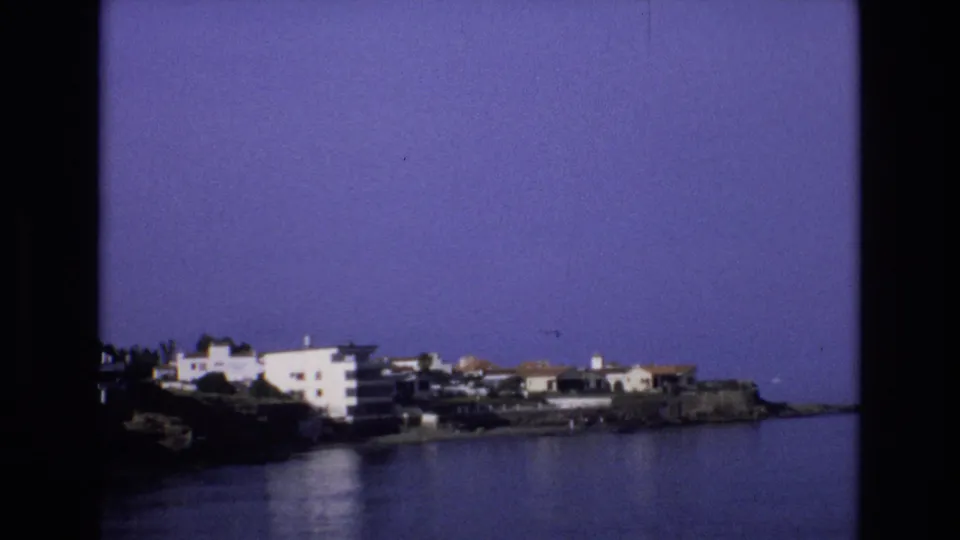 1972 Spain home-movie frame showing white and cream buildings along a rocky shoreline with calm water in the foreground and a clear blue-purple sky above.