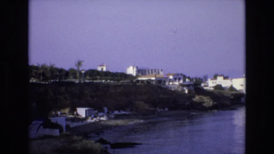 1972 Spain home-movie frame showing a darker rocky shore with palm trees, small waterfront structures, white buildings, and calm water below a clear sky.