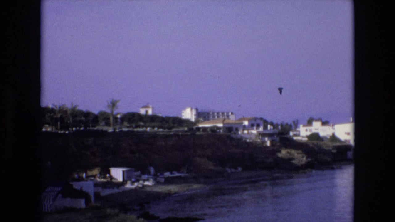 1972 Spain 8mm frame showing a purple-tinted coastal view with white buildings and palm trees above calm water.