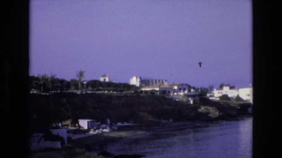 1972 Spain 8mm frame showing a purple-tinted coastal view with white buildings and palm trees above calm water.