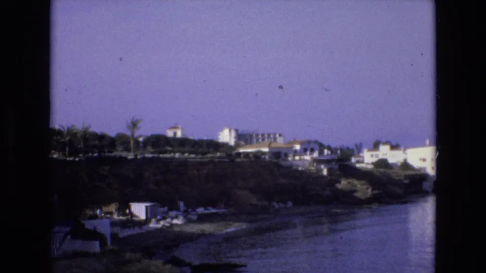 1972 Spain home-movie frame of a coastal hillside settlement seen across calm water, with palms and white buildings visible.