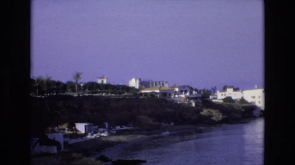 1972 Spain 8mm frame showing white coastal buildings, palm trees, scrub vegetation, and calm water with a soft purple film cast.