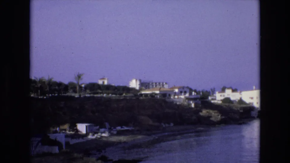 1972 Spain home-movie frame of a quiet shoreline, with white hillside buildings and palms above a broad stretch of water.
