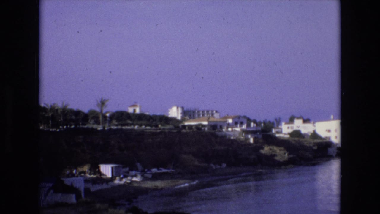 1972 Spain 8mm frame showing a waterfront resort area with hillside buildings, palm trees, beach furniture, and dark film borders.