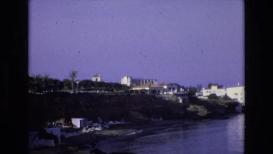 1972 Spain 8mm frame showing a waterfront resort area with hillside buildings, palm trees, beach furniture, and dark film borders.