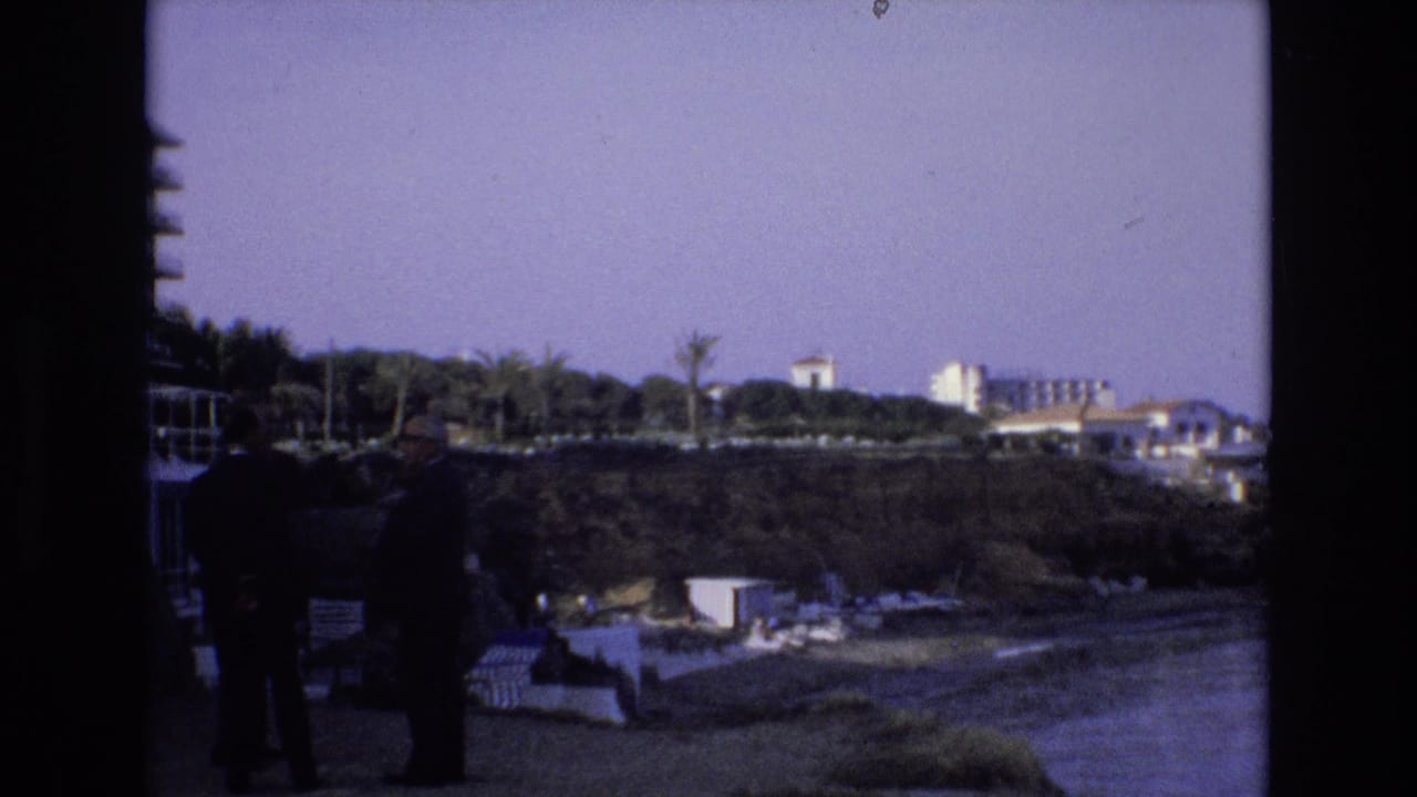 Two men stand near a waterfront overlook with resort buildings beyond.