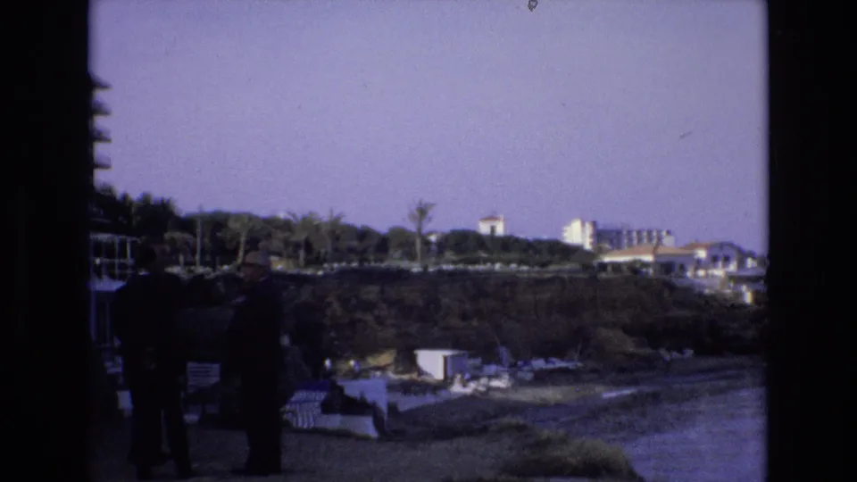 1972 Spain 8mm frame with two suited men in the foreground near a coastal overlook, with palms, beach structures, and resort buildings across the water.