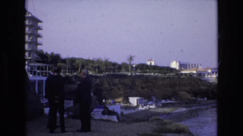 1972 Spain 8mm frame showing two men in dark suits near a waterfront beach area, with a tall building edge, palms, lounge chairs, and hillside resort structures visible.