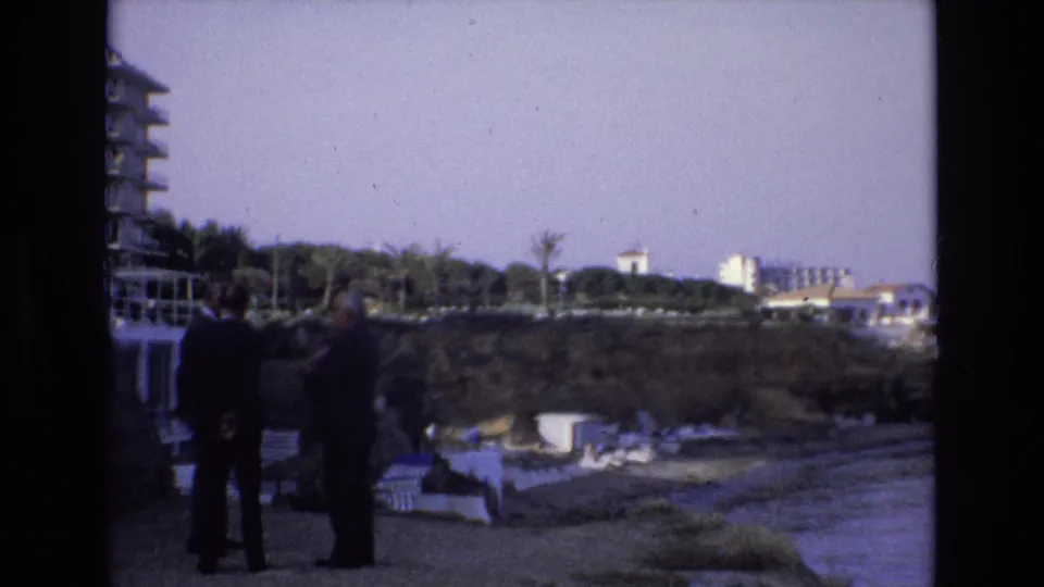1972 Spain 8mm frame of two suited men seen from behind or in profile at a coastal overlook, with beach furniture, palm trees, and low resort buildings in the background.