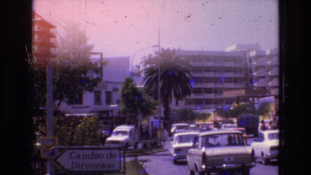 1972 Spain street view with classic cars moving past a palm-lined intersection, multi-story buildings, storefronts, and a visible Cambio de Direccion road sign.