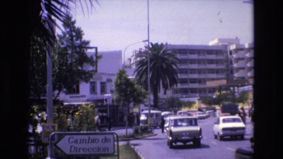 1972 Spain urban traffic scene with a van and compact cars approaching through a palm-lined street, with buildings and the Cambio de Direccion sign in the foreground.