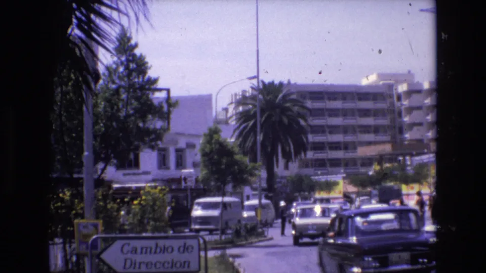 1972 Spain street traffic with a white van, dark sedan, compact cars, palm trees, storefronts, and a Cambio de Direccion sign along the roadside.