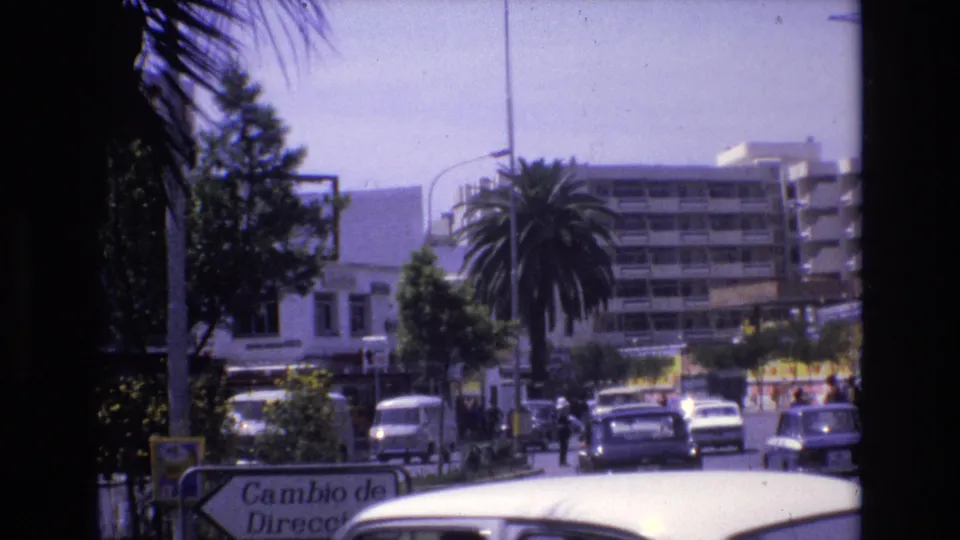 1972 Spain city street view with classic cars, pedestrians, palm trees, a partly visible Cambio de Direccion sign, and multi-story buildings in the background.