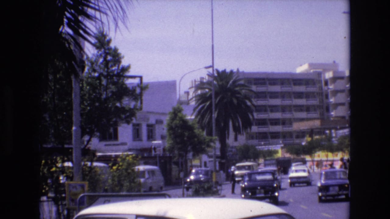 1972 Spain street view with compact cars moving along a sunlit urban road, palm trees, and apartment-like buildings in the distance.