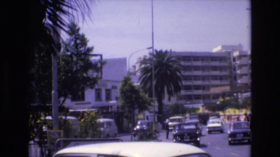1972 Spain street view with compact cars moving along a sunlit urban road, palm trees, and apartment-like buildings in the distance.