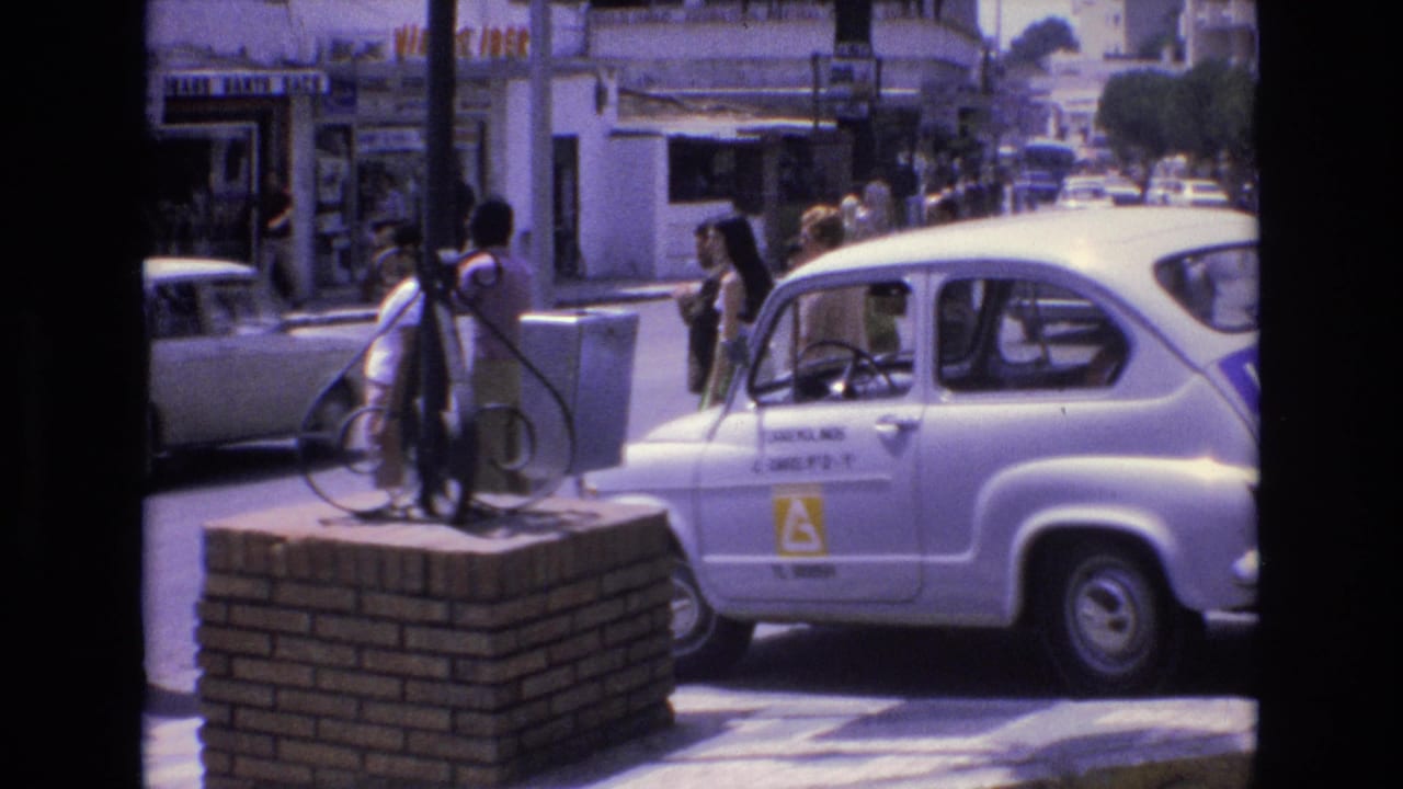 People stand near a white learner-marked car beside a brick platform on a city street.