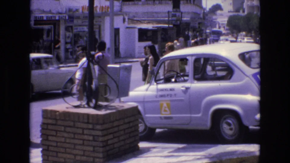 In Spain in 1972, several adults stand near a white learner-marked car parked beside a low brick platform on a busy urban street.