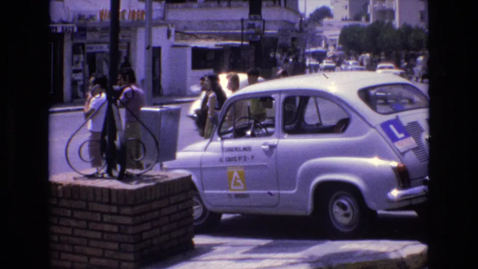 A 1972 Spain street scene frames adults near a white learner-marked car, with other vehicles, storefronts, and sunlit urban traffic in the background.
