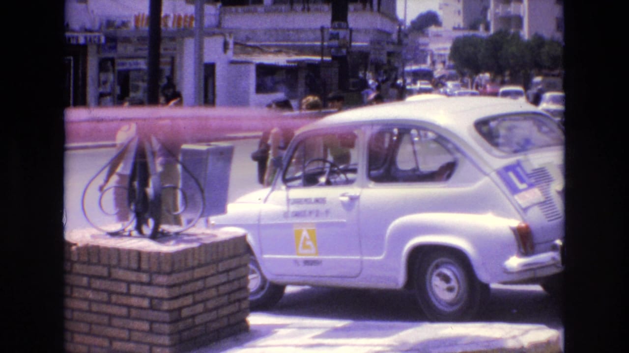 In 1972 Spain, a white learner-marked compact car passes a city sidewalk with pedestrians, storefronts, a brick pillar, and a dark metal post in bright sun.