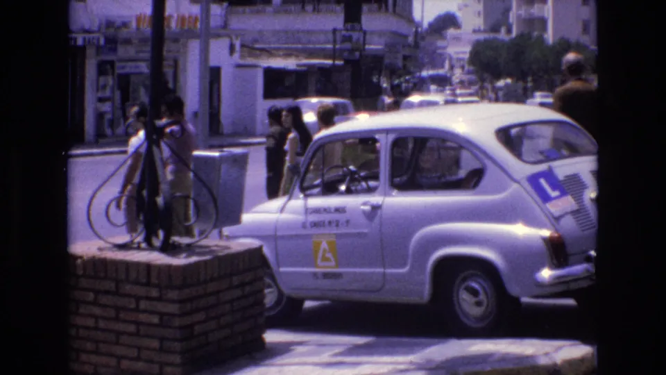 The white learner car is seen from the side on a sunlit Spanish street, with pedestrians standing near the sidewalk and urban buildings in the background.