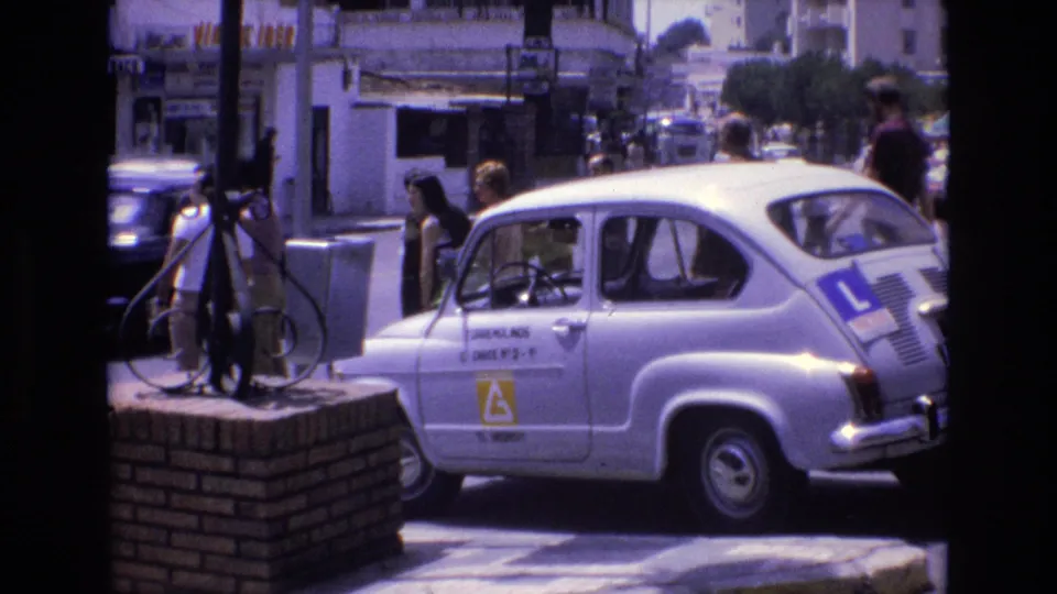 A white learner-marked car continues past the curb in 1972 Spain, framed by a brick pillar, pedestrians, storefronts, and traffic in the background.