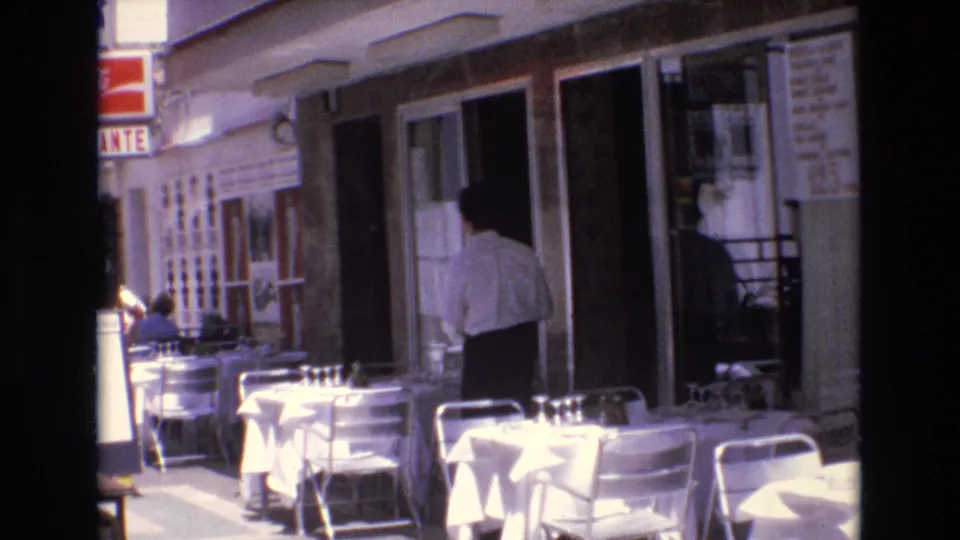 An outdoor restaurant in Spain shows white-clothed tables and metal chairs along the sidewalk, with a man in a light shirt standing near the entrance.