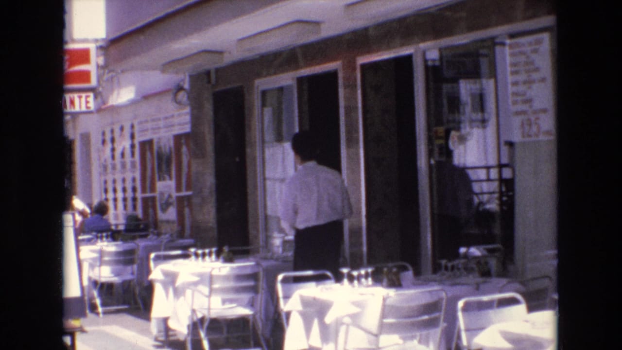 1972 Spain 8mm frame showing white-clothed sidewalk café tables, metal chairs, and a man in a light shirt near a restaurant doorway.