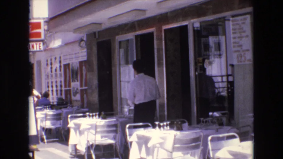 1972 Spain 8mm frame showing white-clothed sidewalk café tables, metal chairs, and a man in a light shirt near a restaurant doorway.