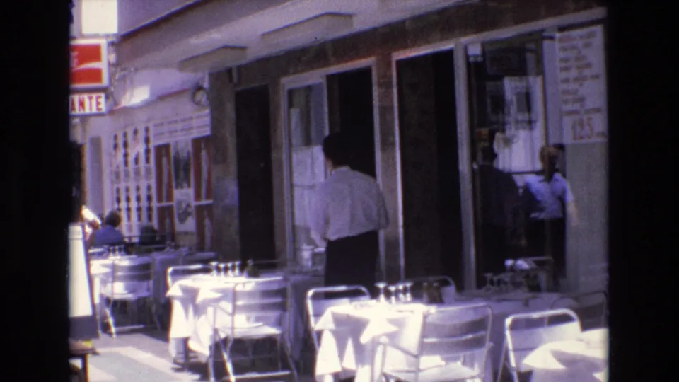 1972 Spain 8mm frame of a sidewalk café with white tables, metal chairs, a man near the entrance, and another person partly visible by the glass door.