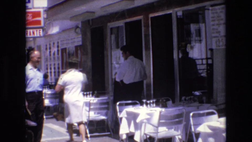 1972 Spain 8mm frame showing a woman in a white dress walking past a restaurant sidewalk café while two men appear near the seating and entrance.