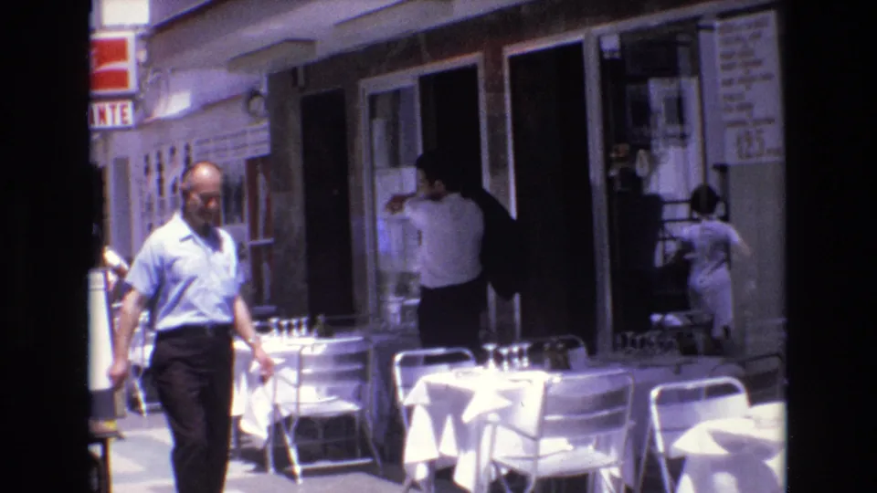 1972 Spain 8mm frame with a man in a blue shirt crossing the foreground, white-clothed sidewalk café tables, and people near a restaurant doorway.