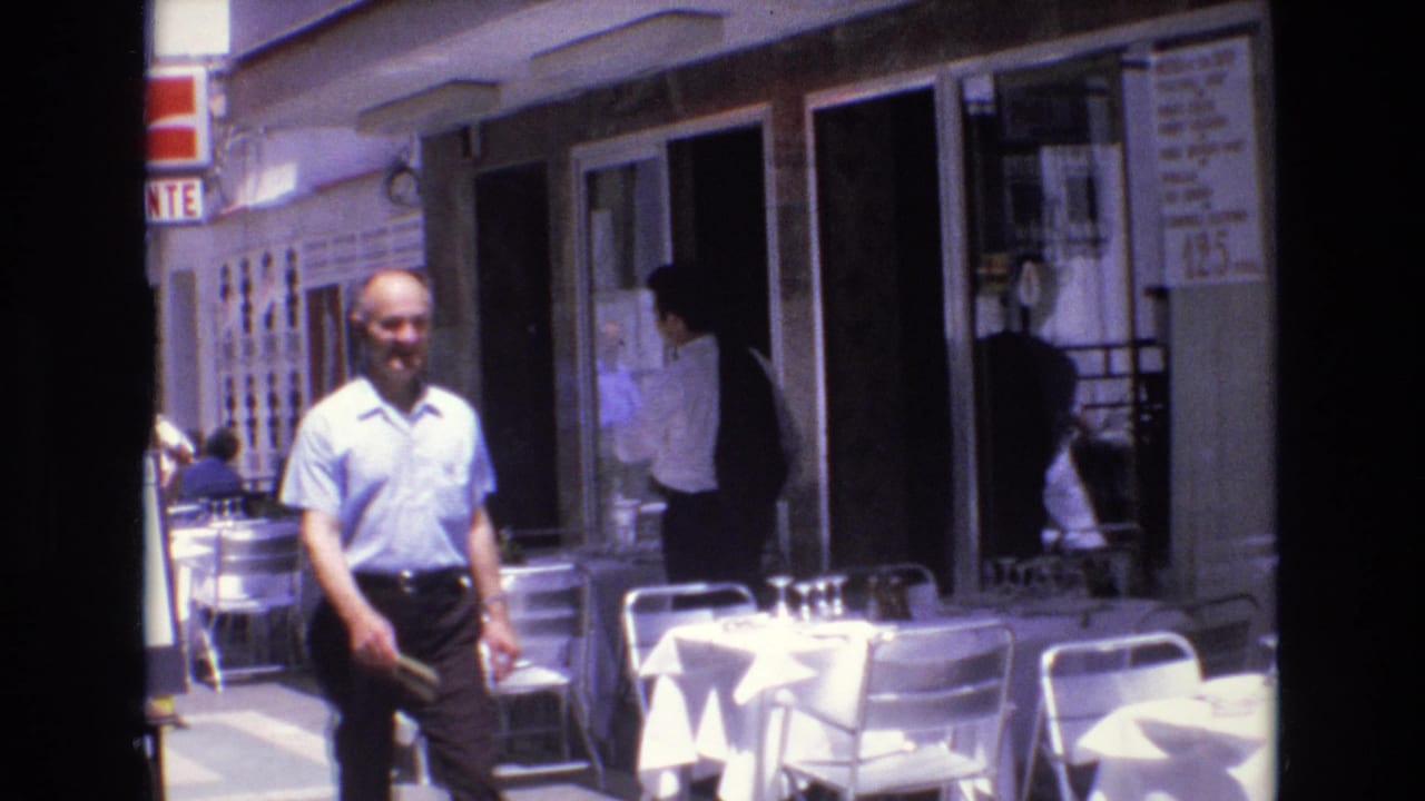1972 Spain 8mm frame showing a man in a light blue shirt walking toward the camera beside white-clothed outdoor cafe tables and metal chairs.