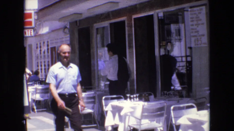 1972 Spain 8mm frame showing a man in a light blue shirt walking toward the camera beside white-clothed outdoor cafe tables and metal chairs.