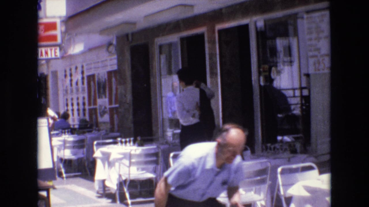 Man bending or moving close to the camera beside outdoor cafe seating.