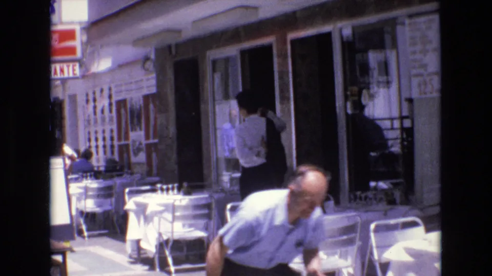 1972 Spain 8mm frame with the man partly close to the camera near cafe chairs, while a staff figure stands by the restaurant entrance behind him.