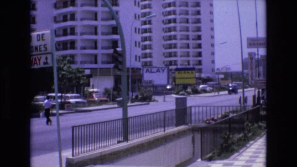 1972 Spain 8mm frame showing a city street view with tall apartment blocks, roadside railings, signs, traffic lights, and cars in bright daylight.