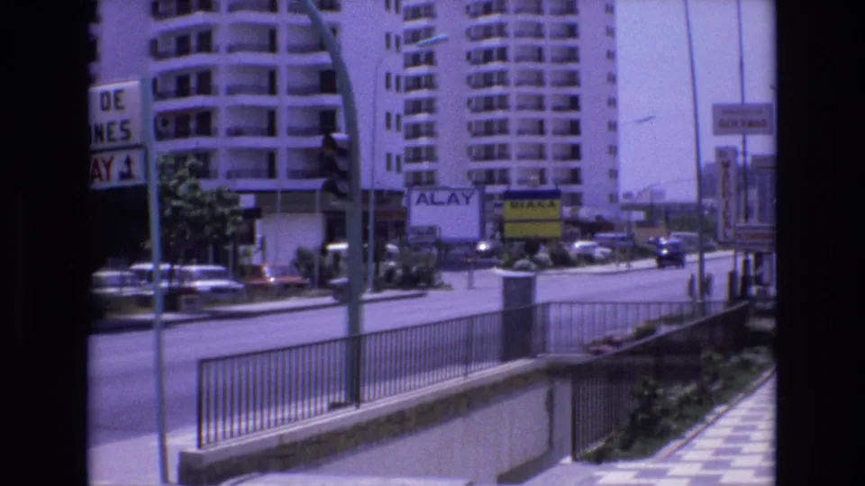 1972 Spain 8mm frame holding on the city street, with apartment buildings, signs, railings, cars, and a sunlit sidewalk visible across the road.