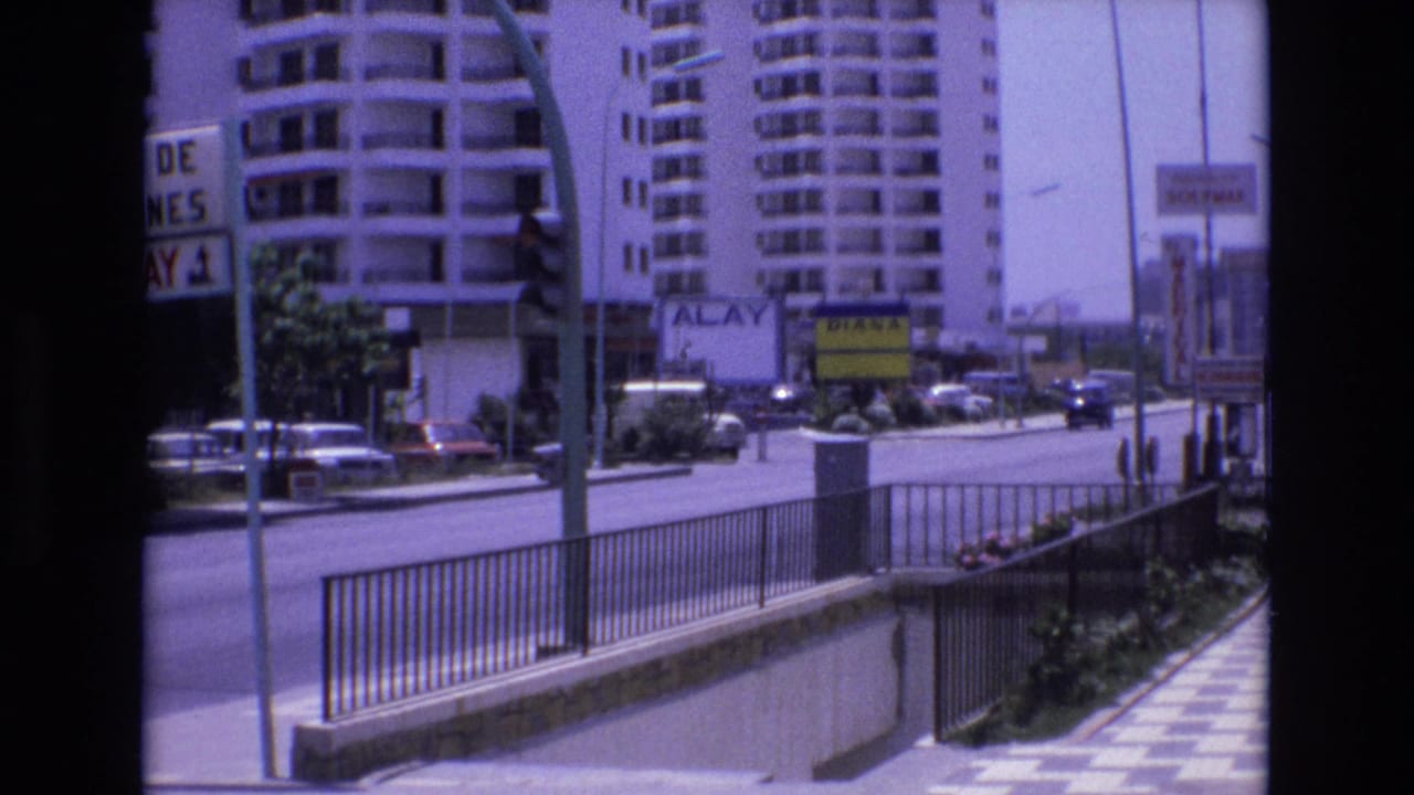 1972 Spain 8mm frame showing a sunny urban street with apartment buildings, metal pedestrian railing, parked cars, road signs, and a stairwell or subway entrance.