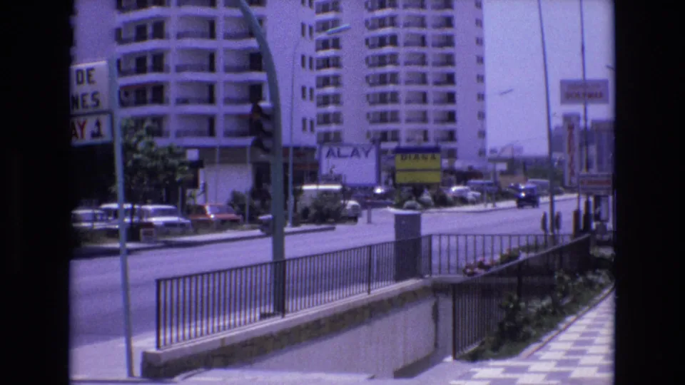 1972 Spain 8mm frame showing a sunny urban street with apartment buildings, metal pedestrian railing, parked cars, road signs, and a stairwell or subway entrance.