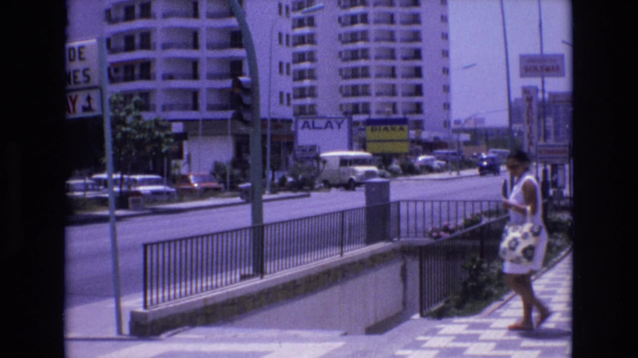 Woman in a white dress walks along a checkered sidewalk beside a railing and city road.