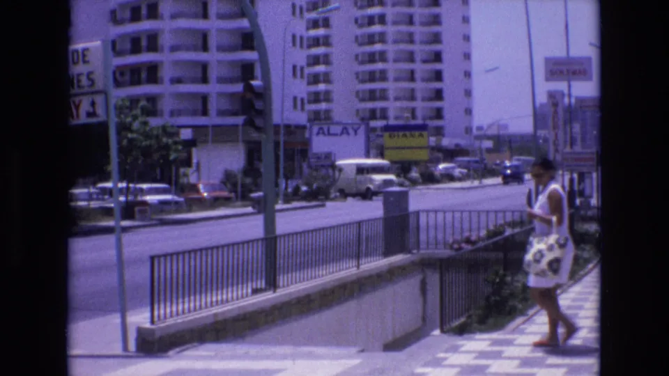 1972 Spain 8mm frame of a woman in a white dress carrying a patterned bag beside a metal railing, with apartment buildings and traffic in the background.