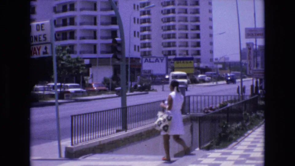 1972 Spain 8mm frame showing a woman in a white dress carrying a patterned bag near a stairwell railing while cars and a delivery van move along the street.