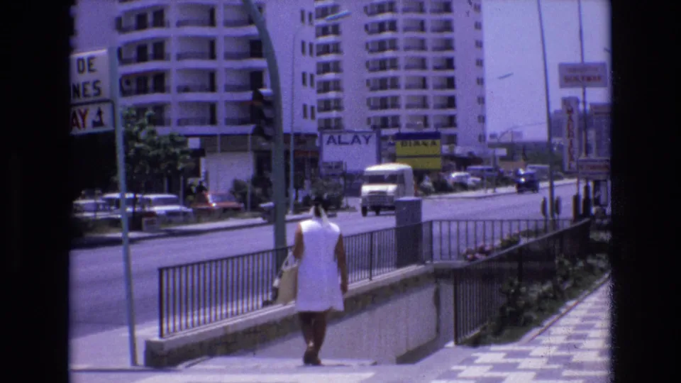 1972 Spain 8mm frame of a woman seen from behind near a stairwell or subway entrance, with a white delivery van, parked cars, apartment buildings, and roadside signs visible.