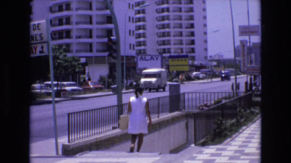 In Spain 1972 home-movie footage, a woman in a white dress descends subway or pedestrian underpass steps beside traffic, railings, high-rise buildings, and visible commercial signage.