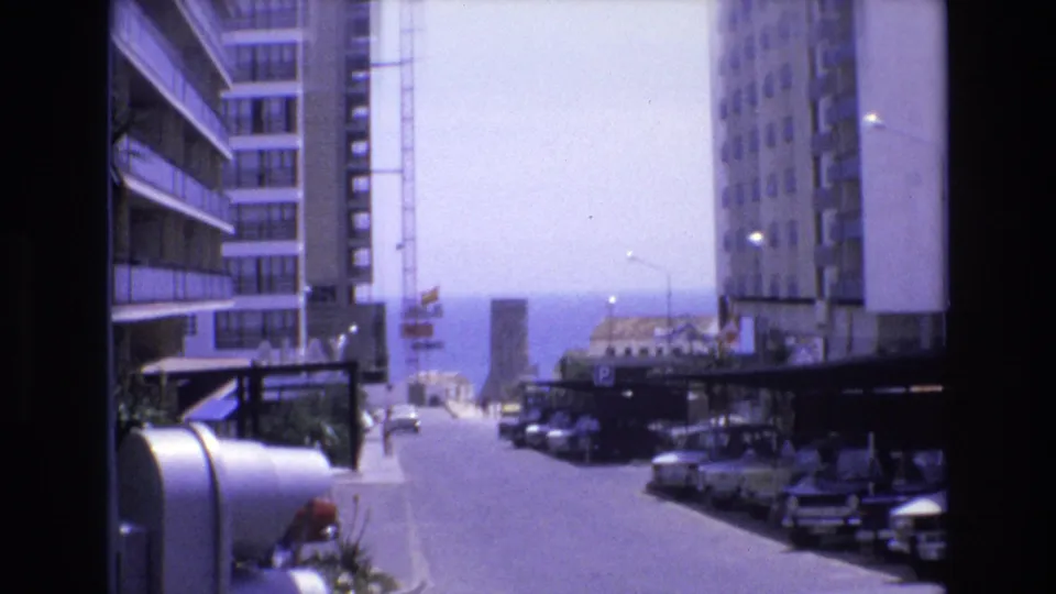The final frame shifts to a 1972 Spain city street perspective between tall buildings, with parked cars along the curb, sidewalk structures, and bright haze at the far end of the road.