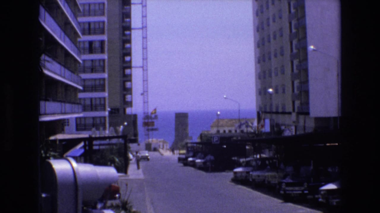 1972 Spain home-movie frame looking down a city street between modern apartment blocks toward the sea, with parked cars, pedestrians, and a crane in the distance.