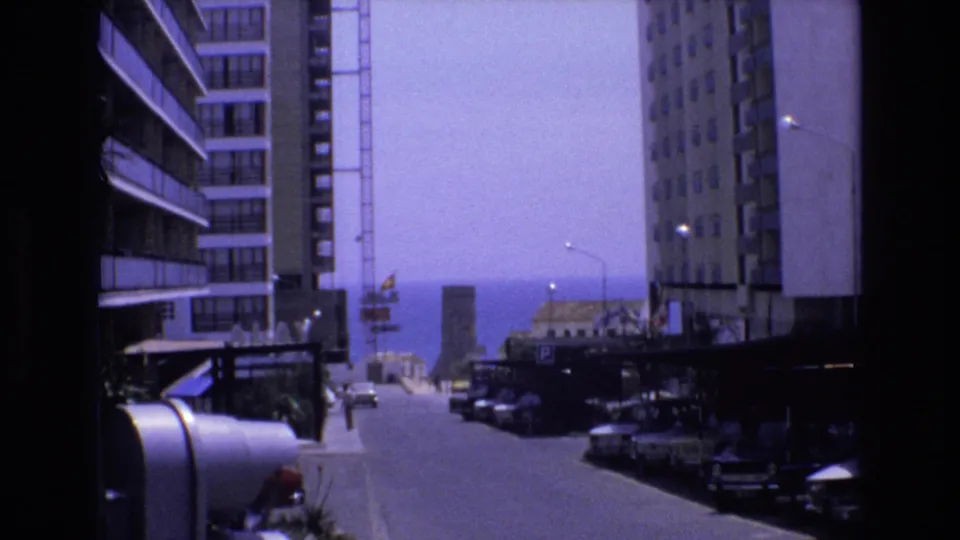 1972 Spain home-movie frame looking down a city street between modern apartment blocks toward the sea, with parked cars, pedestrians, and a crane in the distance.