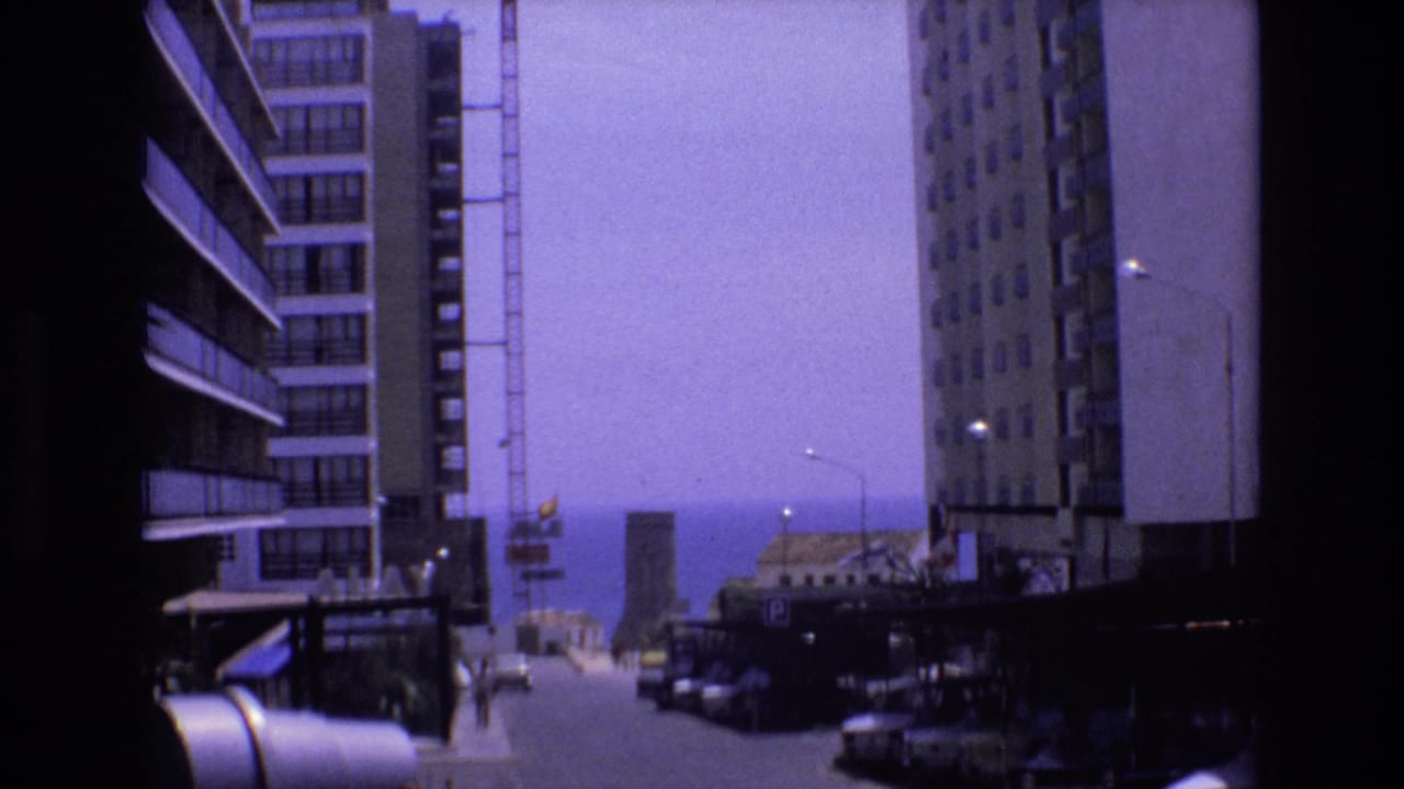 Street scene framed by apartment blocks with parked cars and a distant sea view.