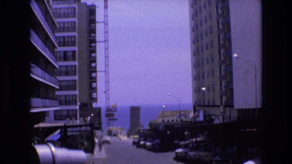 1972 Spain frame showing a sunlit urban street, parked cars along the curb, tall residential buildings, distant pedestrians, and the sea beyond the street.