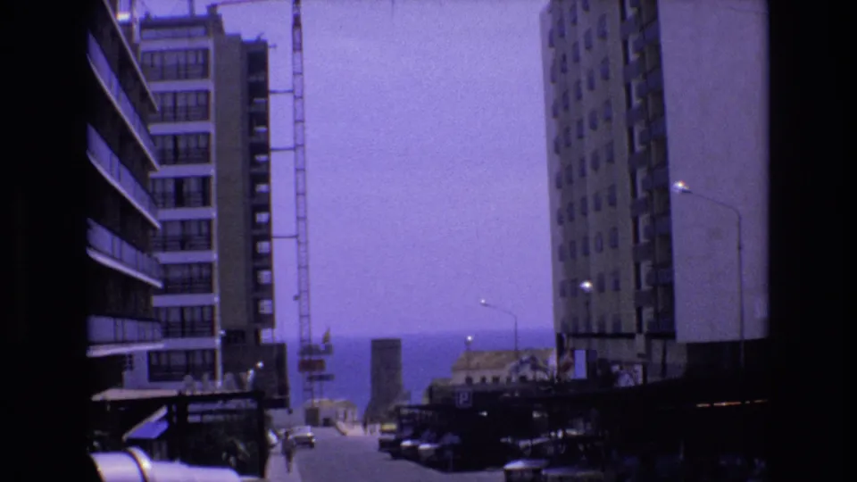1972 Spain home-movie frame with high-rise apartment facades, a sloping street, parked cars, a crane, and a distant sea horizon visible through the buildings.